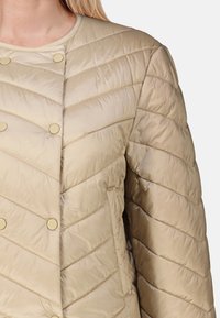 Ljusbeige pufferjacka med kviltat chevronmönster, försedd med snäpplåsförsedda knappar i guldton och rund krage, tillverkad av glänsande tyg.