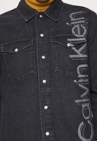 Chaqueta de mezclilla negra con botones plateados, dos bolsillos en el pecho y logo vertical de Calvin Klein en el lado izquierdo, llevada sobre un cuello alto mostaza.