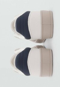 Baskets blanches avec un accent bleu marine sur le talon, présentant un design texturé et une semelle beige. Détails de couture visibles.