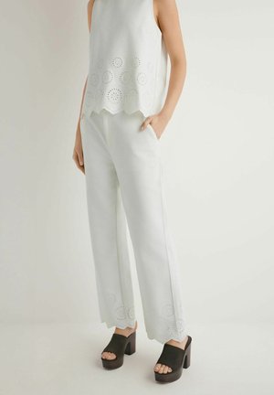 Kobieta w białym, bezrękawnikowym topie i spodniach z wzorami ażurowymi, w połączeniu z czarnymi platformowymi sandałami na otwartym palcu, z rękami w kieszeniach, stoi.