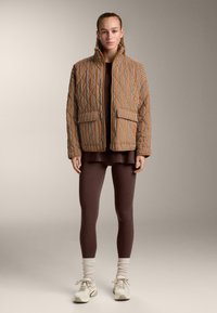 Kobieta ubrana w brązową, w paski puchową kurtkę, ciemnobrązowe legginsy, kremowe skarpetki i białe sportowe buty, stojąca na neutralnym tle.