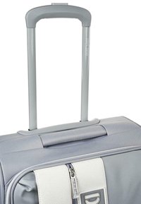 Maleta blanda gris de textura suave, con un asa extensible gris y un bolsillo con cremallera blanca que presenta un detalle de logo.