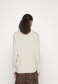Pull beige en maille à coupe décontractée, poignets côtelés et léger effet épaules tombantes, associé à une jupe noire et marron à motifs.