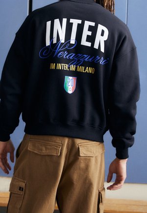 Osoba nosząca czarną bluzę z napisem "INTER Nerazzurri IM INTER, IM MILANO" oraz małym logo w kształcie tarczy w kolorach czerwonym, białym i zielonym na plecach.