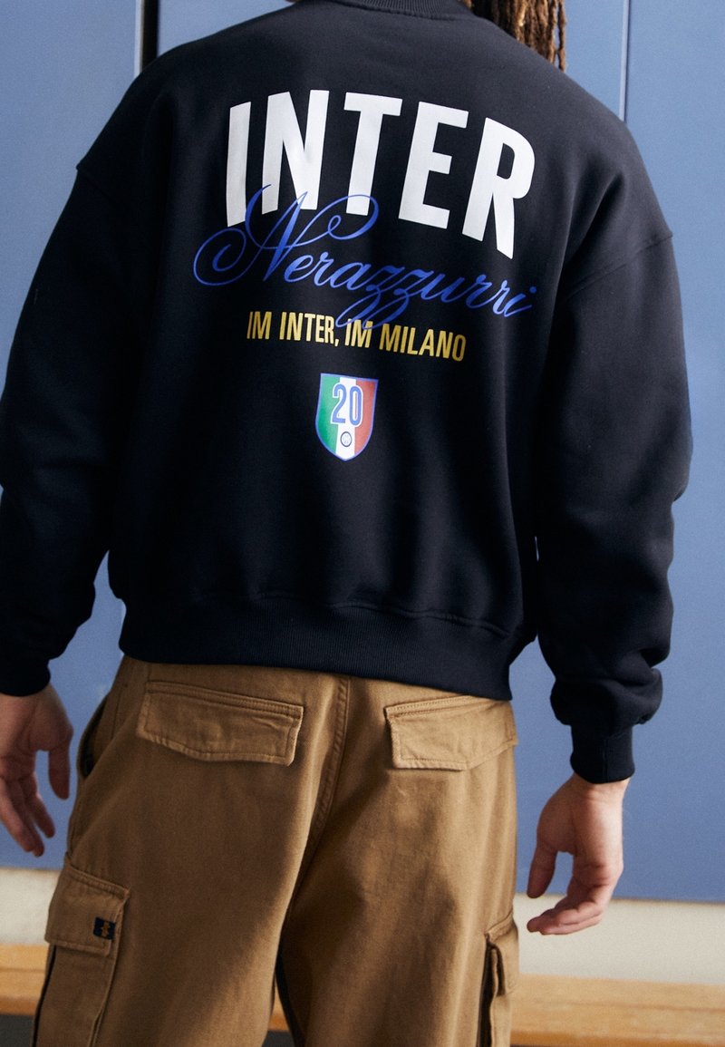 Personne portant un sweat-shirt noir avec le texte « INTER Nerazzurri IM INTER, IM MILANO » et un petit logo en forme de bouclier en rouge, blanc et vert dans le dos.