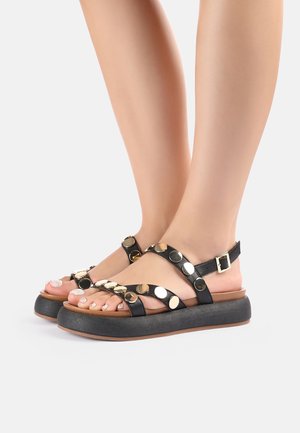 Voeten met zwarte plateauzandalen met gouden, ronde studs en enkellinten, tegen een eenvoudige witte achtergrond.
