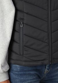 Gilet noir isolé avec un motif en chevrons, comportant une poche zippée sur le côté gauche et une chemise à manches longues grise en dessous.