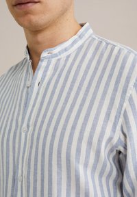 Camicia di lino a righe con linee orizzontali alternate di azzurro chiaro e bianco, dotata di colletto a banda e patta frontale con bottoni.