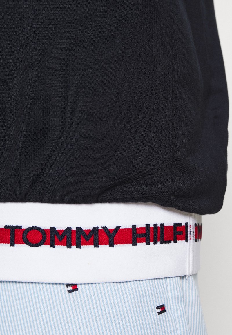 Tessuto nero con fascia elastica bianca caratterizzata da una striscia rossa e testo "TOMMY HILFIGER" in nero sopra pantaloni a righe azzurro chiaro con piccoli loghi a bandiera.