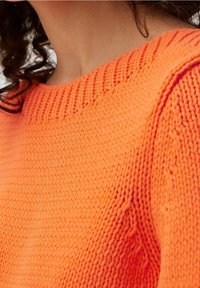 Primo piano di una persona che indossa un luminoso maglione lavorato a maglia arancione con scollatura rotonda e motivo a punto testurizzato.