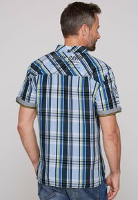 Camicia a maniche corte a quadretti in blu, verde e nero. Presenta un colletto con bottoni, un bordo giallo a contrasto e il logo del marchio sulla schiena.