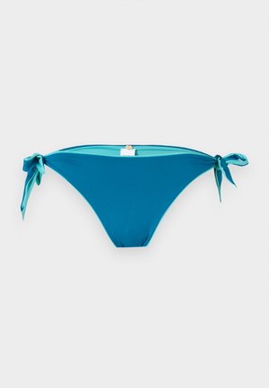 Femme debout portant un haut de bikini blanc, un bas de bikini bleu avec des liens latéraux, et des sandales noires sur un fond clair uni.