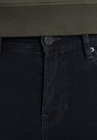 Jeans en denim bleu marine avec une finition texturée. Présentent une fermeture à bouton et des poches, partiellement couvertes par un sweat à col ribbed vert olive.