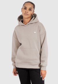 Haut à capuche beige clair avec une poche avant, un cordon de serrage au niveau de la capuche et un logo discret sur la poitrine. Fabriqué en tissu doux et texturé. Coupe décontractée.
