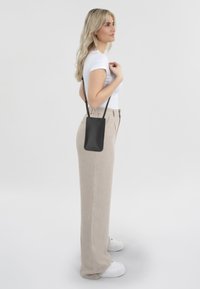 Petit sac en cuir noir à bandoulière avec un design élégant, porté sur le côté. Modèle en haut blanc ajusté et pantalon beige taille haute.