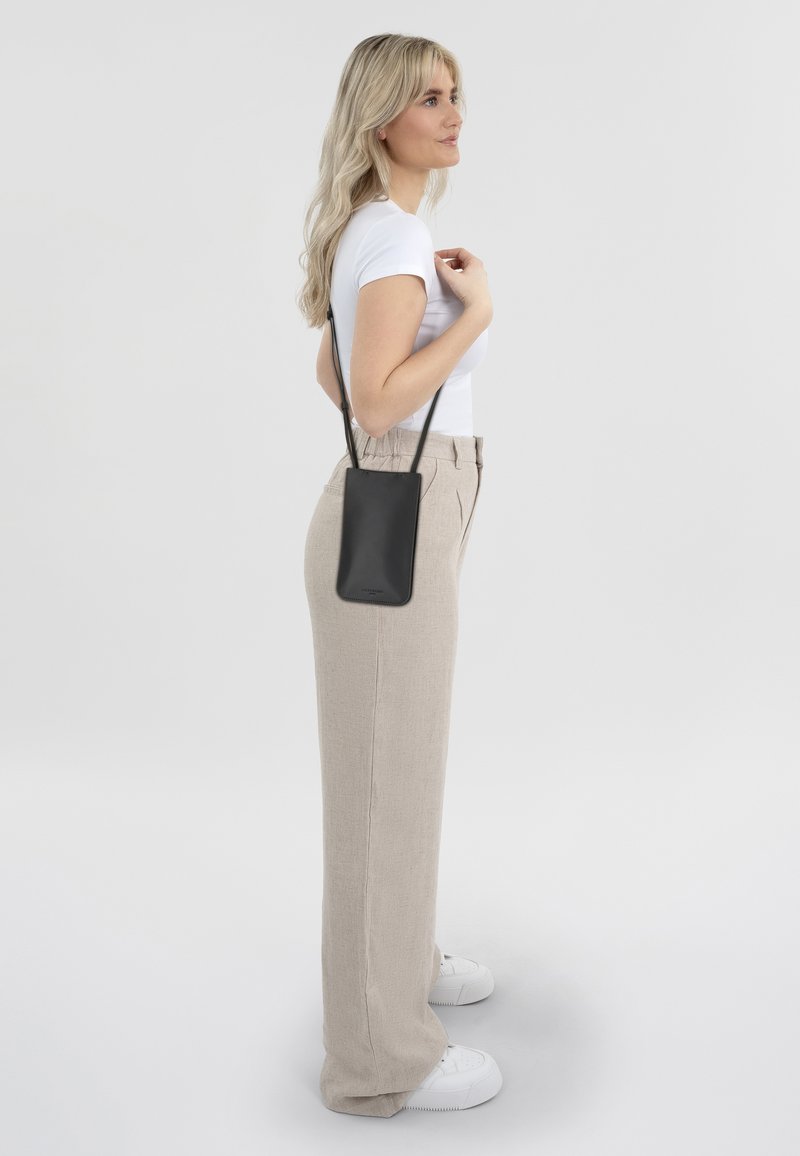 Petit sac en cuir noir à bandoulière avec un design élégant, porté sur le côté. Modèle en haut blanc ajusté et pantalon beige taille haute.