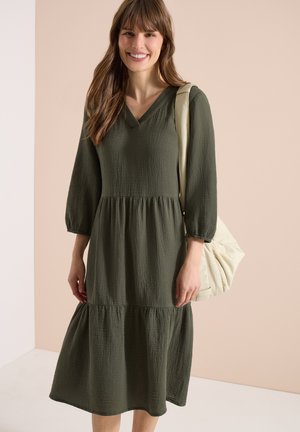 Jeune femme portant une robe midi olive à manches longues avec un col en V, portant un sac à bandoulière crème souple, souriant devant un fond beige.
