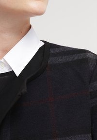 Schwarz-weiß kariertes Sweatshirt aus Wolle mit runder Ausschnitt und einem kontrastierenden schwarzen Saum. Weißes Hemd mit Kragen darunter.