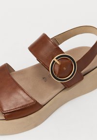 Brown leather sandal with a wide strap and gold and black circular buckle detail, featuring a beige platform sole and soft interior lining.