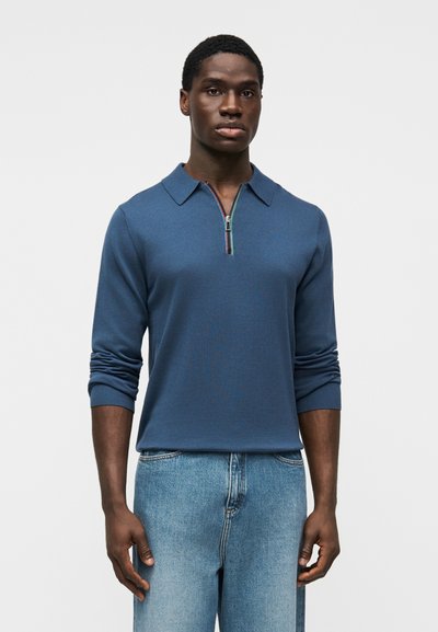 Homme portant un polo bleu à manches longues avec fermeture éclair et col, ainsi qu'un jean bleu clair, debout devant un fond blanc uni.