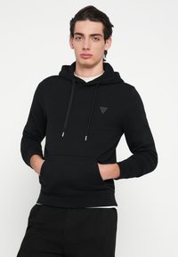 Μαύρη φούτερ με κουκούλα από απαλό υλικό, με μπροστινή τσέπη, κορδόνι στην κουκούλα και διακριτική μπαλώματος με λογότυπο στο στήθος.