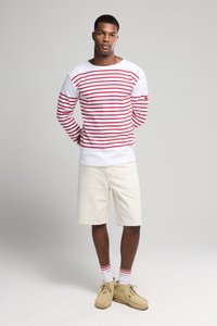 Hvid langærmet skjorte med røde striber, parret med kremfarvede shorts. Beige sko og hvide sokker med røde accenter fuldender outfittet.