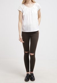 Haut blanc à manches courtes avec un accent en dentelle, associé à un jean skinny marron foncé avec des déchirures aux genoux, et des chaussures noires à lacets.