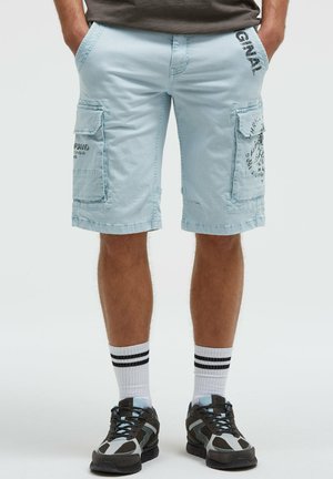 Mann trägt hellblaue Cargo-Shorts mit Seitentaschen, schwarz-weiße Sneakers und weiße Socken mit zwei schwarzen Streifen, steht mit den Händen in den Taschen.