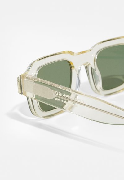 Gafas de sol de plástico transparente con lentes verdes, forma angular, montura transparente y logo grabado en la parte interior de la patilla.