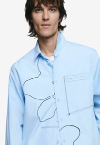 Hele sinine pikkade varrukatega särk, millel on rinnatasku ja esiküljel julge must joone muster. Sile materjal, nööbitav stiil.