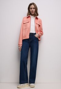 Broskvová bomber bunda se dvěma předními kapsami, kombinovaná s tmavě modrými rozšířenými džínami s vysokým pasem a béžovými ležérními botami.