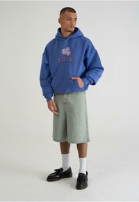 Blå hættetrøje med trykt logo "Y.D.F.C." i rød, parret med lyse denimshorts og sorte sko, båret med hvide sokker.