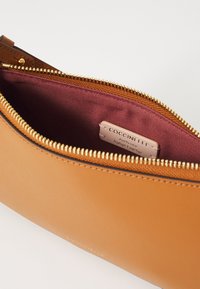 Pochette zippée en cuir marron clair ouverte, montrant une doublure en tissu bordeaux et une étiquette cousue portant l'inscription "Coccinelle Cuir Italien Authentique".