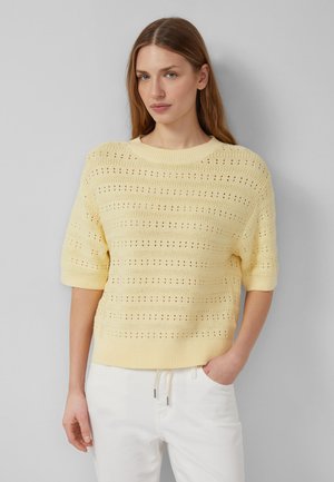 Donna con capelli lunghi che indossa un maglione a maniche corte in maglia giallo chiaro e pantaloni bianchi, in piedi davanti a una parete grigio chiaro con una mano in tasca.