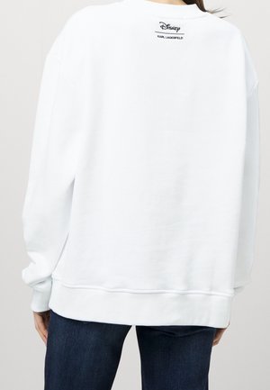 Osoba ubrana w biały sweter z logo "Disney Karl Lagerfeld" na górnej części pleców, w połączeniu z ciemnoniebieskimi dżinsami.