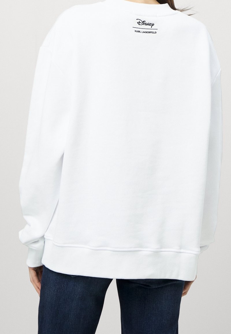 Personne portant un sweat-shirt blanc avec le logo "Disney Karl Lagerfeld" dans le haut du dos, associé à un jean bleu foncé.