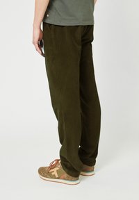 Olijfgroene corduroybroek met een relaxte pasvorm, voorzien van subtiele verticale ribbels en een elastische tailleband; gecombineerd met bruine schoenen.