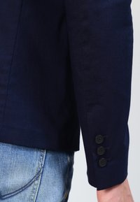Navy blue fabric jacket sleeve with three black buttons and a clean seam at the cuff, paired with light blue denim.