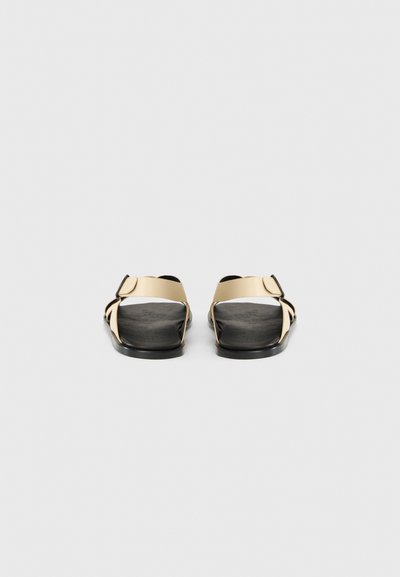 Sandali neri con design a fascia incrociata larga in materiale sintetico beige. Plantare texturizzato e suole piatte. Dettaglio semplice con fibbia sulle fasce.