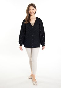 Cardigan nero a maglia con scollo a V, chiusura con bottoni e motivi testurizzati. Abbinato a pantaloni beige aderenti e scarpe di colore chiaro.