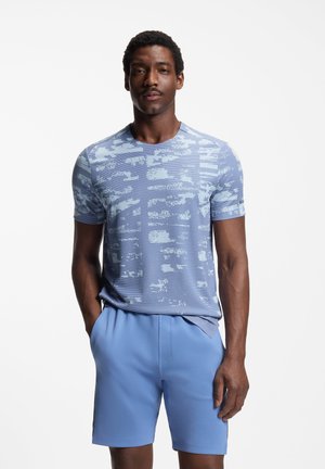 Mężczyzna stojący z rękami w kieszeniach, ubrany w jasnoniebieską koszulę z krótkim rękawem w wzory oraz jednolite jasnoniebieskie szorty na białym tle.