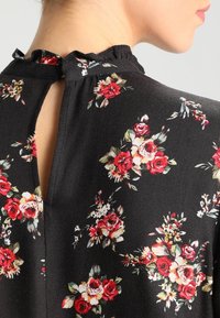 Blouse noire à motifs floraux, avec un dos à trou de serrure, un col à volants et des motifs de roses multicolores. Tissu doux avec un léger éclat.