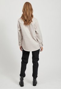 Lys beige langærmet skjorte med afrundet kant, rygskuldersøm detalje, parret med sorte tapered bukser og sorte snørebåndstøvler.