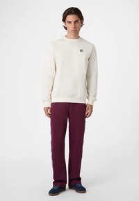 Crèmekleurige sweatshirt met geribbelde manchetten, bordeauxrode trainingsbroek met zijstrepen, en marineblauwe sneakers met geaccentueerde textuur.