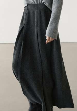 Femme portant une jupe volumineuse gris foncé avec des plis et un pull à manches longues gris clair, debout à l'intérieur sur un sol clair.