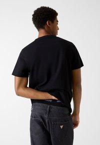 Czarna bawełniana koszulka z krótkim rękawem, widok z tyłu. Noszona z ciemnoszarymi dżinsami z widocznym pasem i trójkątnym logo na kieszeni.