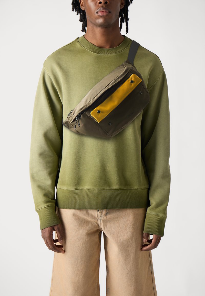 Mochila de cintura de color verde oliva con un panel frontal amarillo, con cremalleras y herrajes negros. Lleva puesta sobre un sudadera verde a juego.