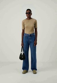 Beige eng anliegendes T-Shirt, hoch taillierte blaue Jeans mit weitem Bein, grüne Slipper, schwarze Sonnenbrille und eine kleine schwarze Handtasche.