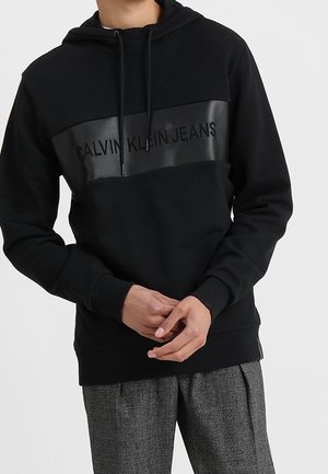 Schwarzer Hoodie mit Kordeln, mit einem horizontalen glänzenden Panel mit dem Text "CALVIN KLEIN JEANS", kombiniert mit grauer gemusterter Hose.