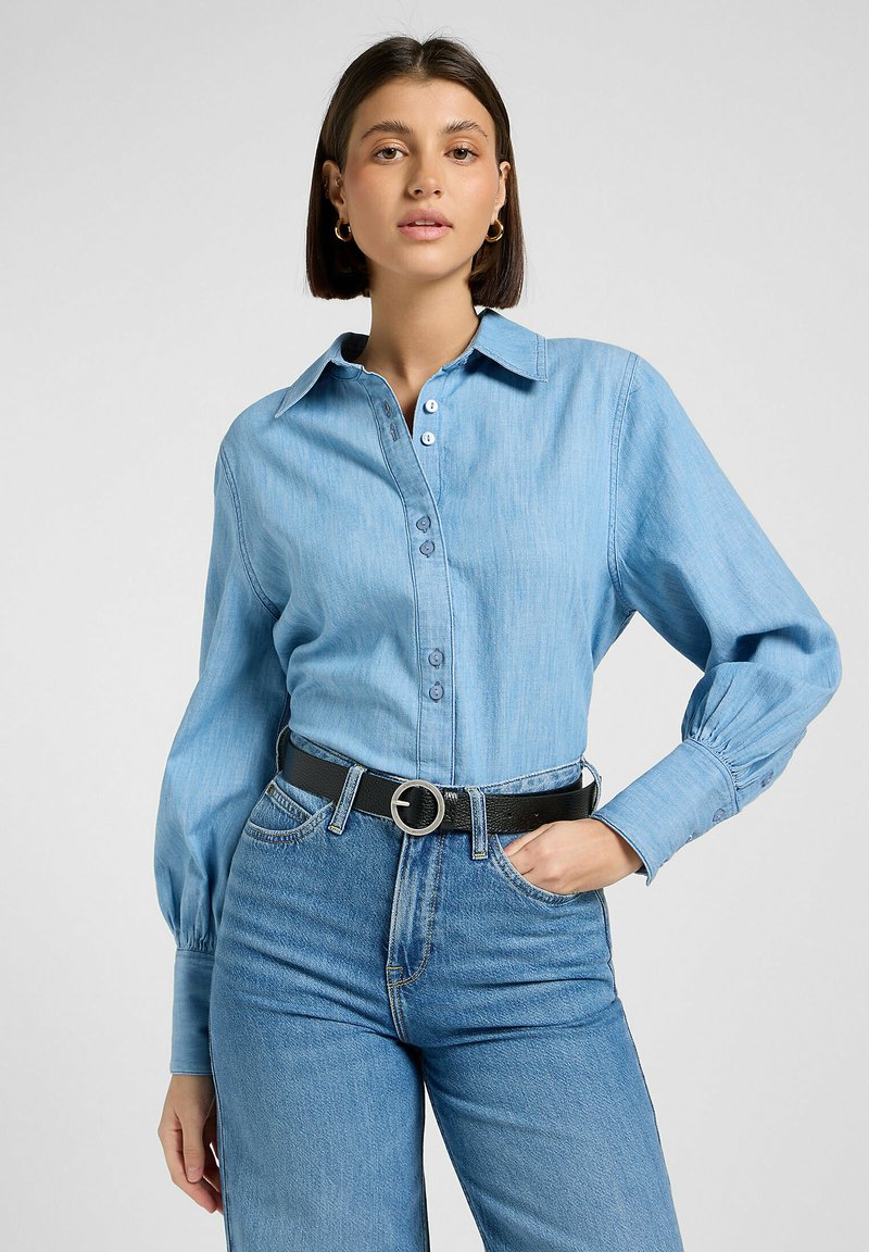 Camisa de mezclilla azul claro con mangas largas, con botones en la parte delantera y un cuello, combinada con jeans azules de tiro alto y un cinturón negro con hebilla circular.